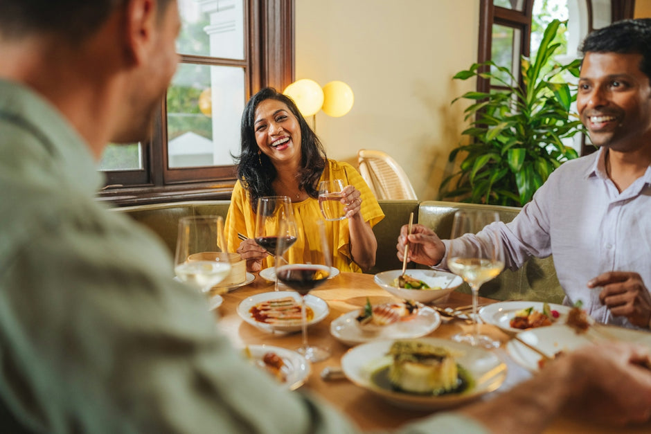 Three people laughing while dining at a restaurant table with wine and food